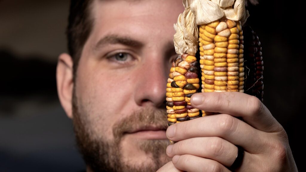 Headshot of Roo George-Warren, who holds an ear of Catawba corn obscuring his right eye.