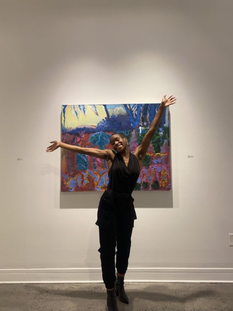 Marquia standing in front of one of her paintings with her arms raised.