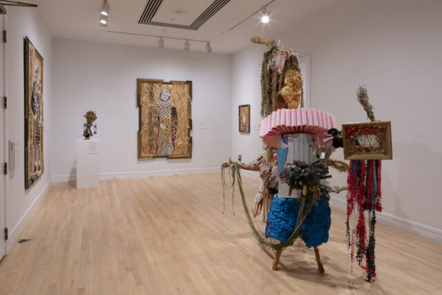 Gallery installation with two large embellished portrait works on plywood, a small bust-like sculpture on a pedestal, and a vibrant mixed-media freestanding sculpture with pink, blue, and red elements in the foreground.