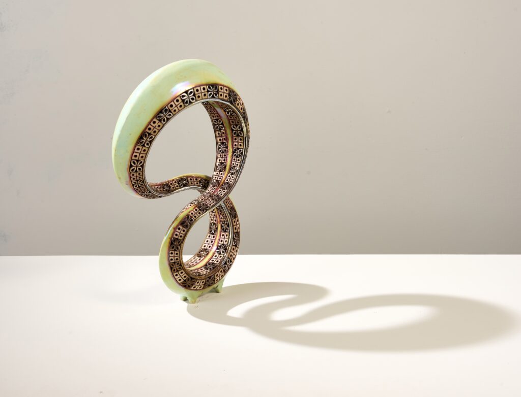 A sculptural artwork featuring two intertwined, ribbon-like loops forming an abstract figure-eight shape. The outer edges are smooth and light green, while the inner surfaces display intricate geometric patterns in bronze and copper tones. The sculpture casts a distinct shadow on the white surface beneath it, enhancing the sense of depth.