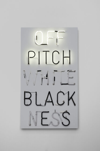Neon artwork with vertically stacked words: 'OFF', 'PITCH', 'WHITE', 'BLACKNESS'. The word 'Next' appears inside the 'O' of 'OFF'. Some letters are illuminated in various colors, others are unlit