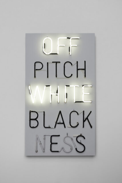 Rectangular panel with neon lights spelling 'OFF PITCH WHITE BLACKNESS'. 'OFF' and 'WHITE' glow in white neon; 'PITCH', 'BLACK', and 'NESS' are in unlit black tubing against a light gray background.