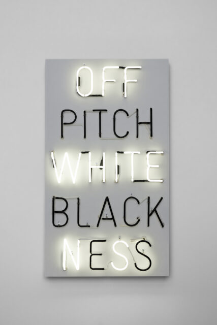 Rectangular neon sign displaying the words 'OFF PITCH WHITE BLACKNESS'. 'OFF', 'WHITE', and 'NESS' are in white neon; 'PITCH' and 'BLACK' are in black neon.