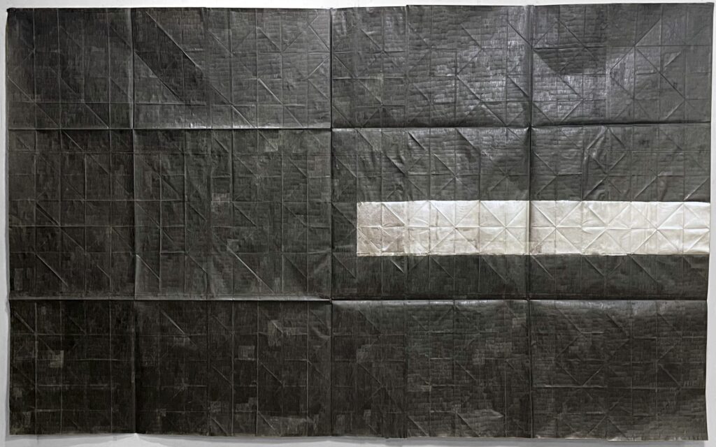 A large, rectangular monochrome artwork composed of heavily creased paper sheets arranged in a grid pattern. The surface is predominantly dark charcoal grey or black with visible grid lines and diagonal folds. A single horizontal band of lighter, off-white or silver paper sheets extends across the middle right portion of the composition.
