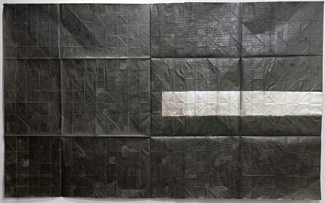 A large, rectangular monochrome artwork composed of heavily creased paper sheets arranged in a grid pattern. The surface is predominantly dark charcoal grey or black with visible grid lines and diagonal folds. A single horizontal band of lighter, off-white or silver paper sheets extends across the middle right portion of the composition.