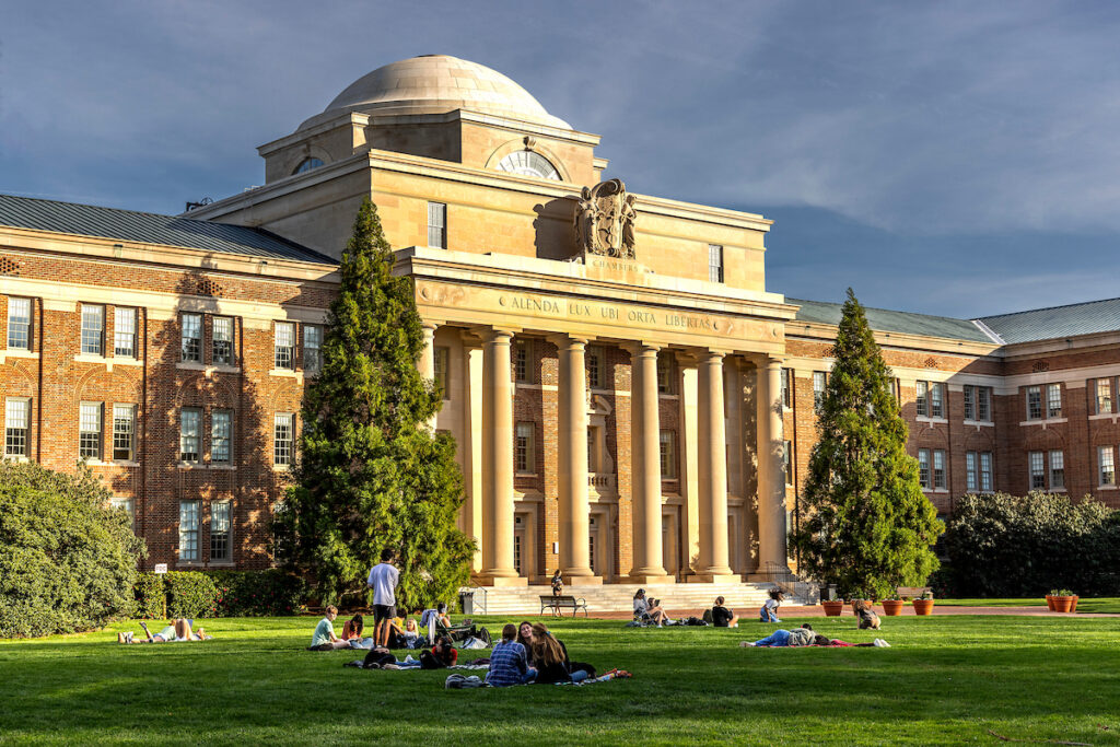 A large, symmetrical brick building with classical architectural details features a central stone portico with large columns supporting a lintel inscribed with "ALENDA LUX UBI ORTA LIBERTAS" and "CHAMBERS." A prominent dome sits atop the central structure. In the foreground, people are scattered across a grassy lawn, some sitting on blankets or lounging, under a partly cloudy sky.