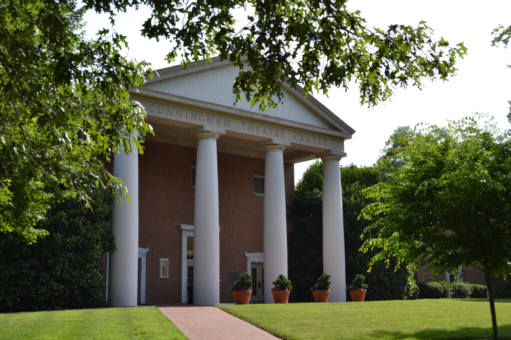 A brick building features a prominent white classical portico with four large columns supporting a pediment. The text "CUNNINGHAM THEATRE CENTER" is inscribed along the architrave above the columns. A brick pathway leads toward the entrance, which is flanked by potted plants, while green lawns, trees, and dense shrubbery frame the structure.