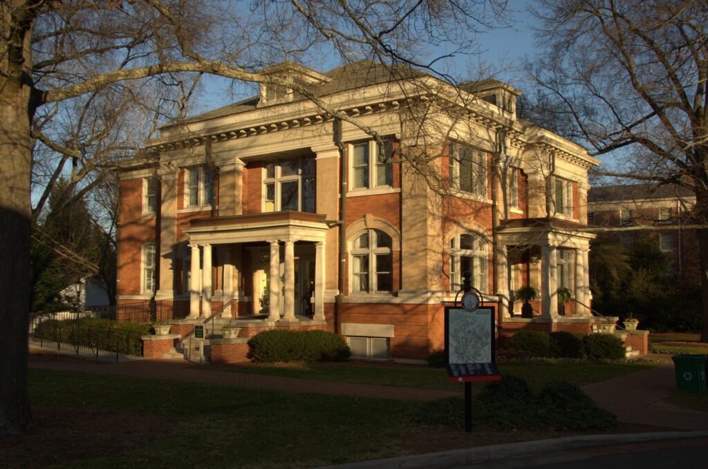A multi-story brick and stone building featuring classical architectural elements, columns at the entrance porches, decorative cornices, and arched windows. The building is framed by bare tree branches in the foreground and a lawn with a map sign stands out front.