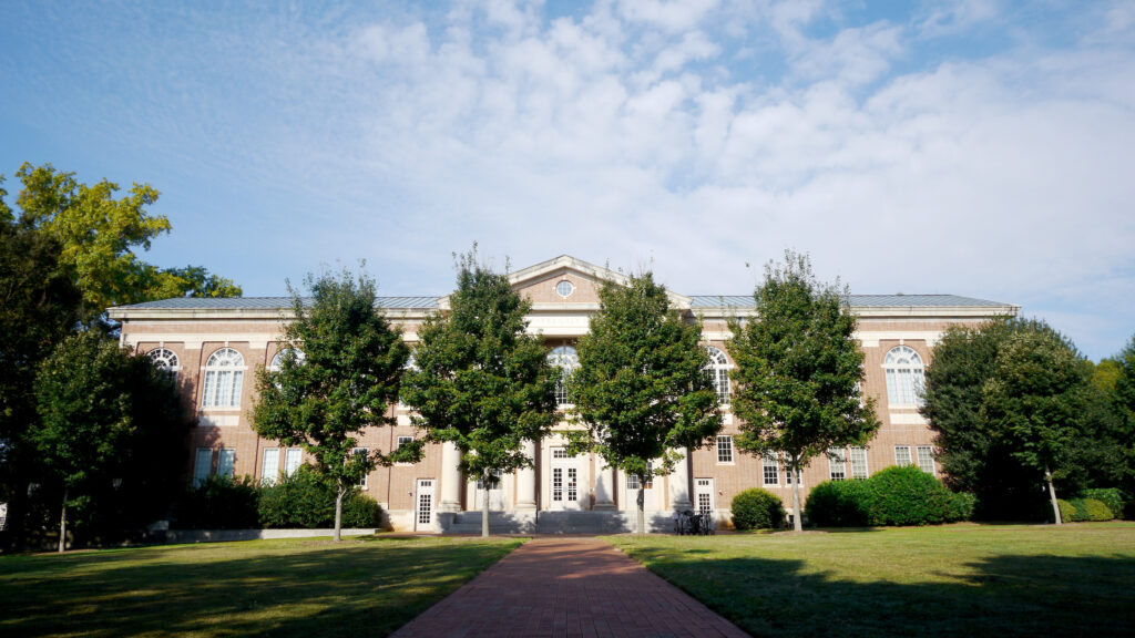 A symmetrical brick building features a prominent central section with columns and a gabled pediment containing a small circular window. The facade is lined with arched windows on the upper level and rectangular windows below. A straight brick path leads directly toward the central entrance from the foreground, flanked by green lawns and a row of mature deciduous trees that partially obscure the front of the building. The sky above is blue with light, scattered clouds.