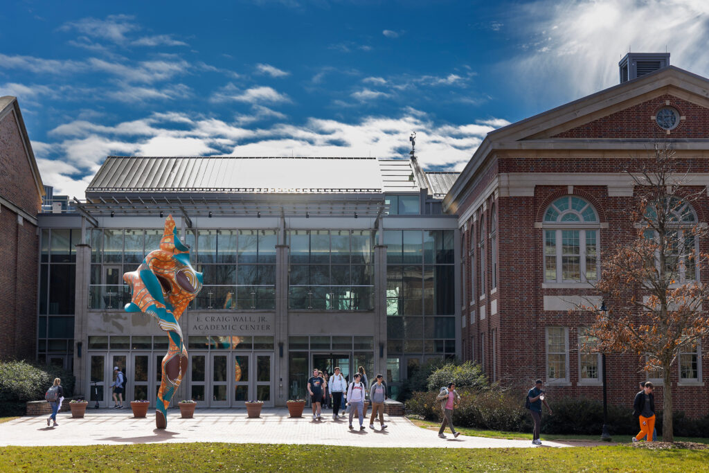 A modern brick and glass building features a large glass entryway with the inscription "E. CRAIG WALL, JR. ACADEMIC CENTER." To the left of the entrance stands a tall, abstract, multi-colored sculpture with swirling orange, blue, and brown patterns. Several people are walking on the paved plaza in front of the building, which is bordered by a grassy lawn under a blue sky with scattered clouds.